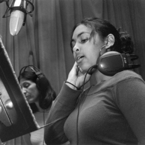 Black and white photo of a student speaking into a microphone, wearing headphones. A second person wearing headphones in the background.
