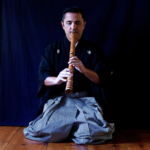 Kodi Araki the sixth kneels on a wooden floor, playing a wooden flute.