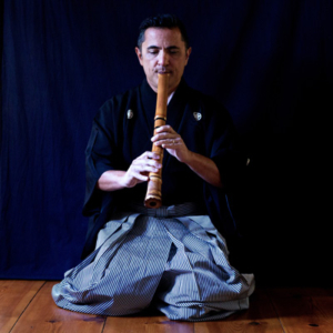 Kodi Araki the sixth kneels on a wooden floor, playing a wooden flute.
