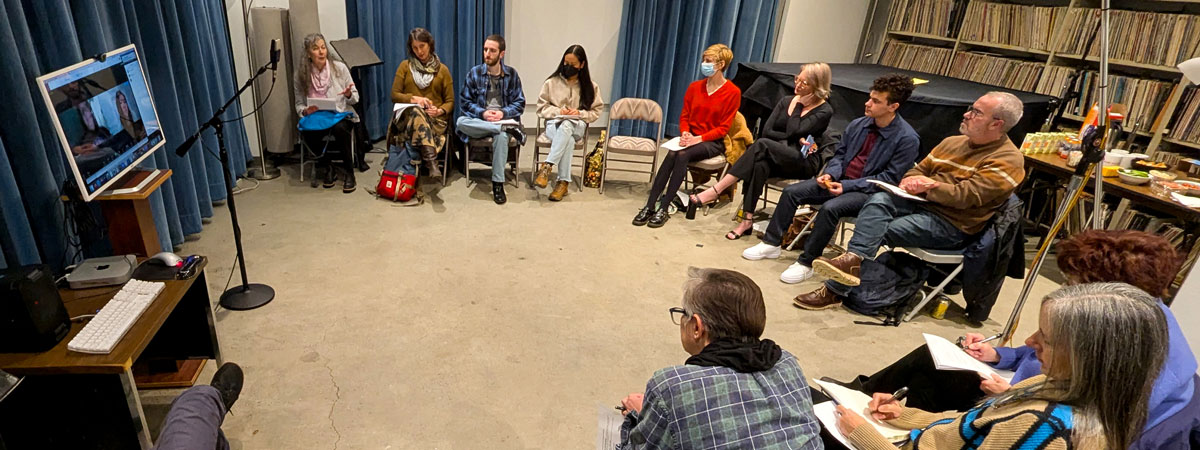 A group of people sitting in a circle in a recording studio. A computer screen on the left shows two people in a zoom meeting.