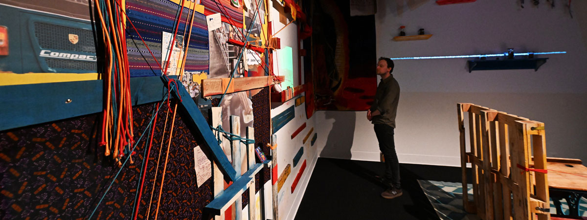 A person stands in a gallery space looking at a wall covered with fabric, string, and other materials. A structure made of wood pallets is behind him.