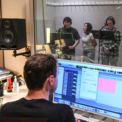 In the foreground, a person at a computer screen showing an audio recording session. Through a glass window, three people in a recording studio. Two wear headphones and stand at microphones and music stands.
