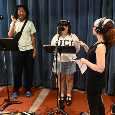 Three people in a recording studio, wearing headphones. Two stand at microphones and music stands.