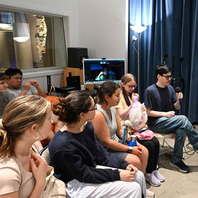 People sitting in a room with concrete floors and blue curtains. One holds a microphone.