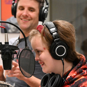 A smiling young person wears headphones at a microphone, while someone in the background, also wearing headphones, looks on and smiles.