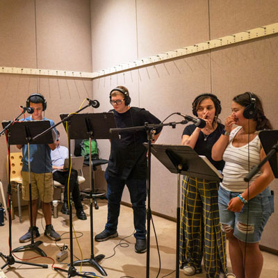Four people in a recording studio, wearing headphones and standing at microphones and music stands.