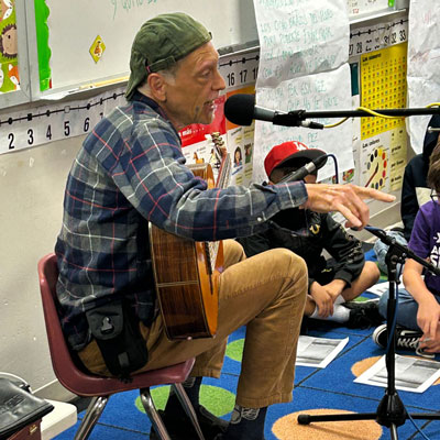 Abel Rocha, in an elementary school class room, sits with a guitar and speaks into a microphone. Students sit on the floor listening.