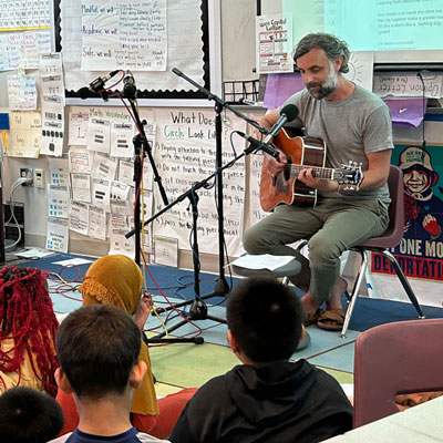 Tito Ramsey sits in an elementary school classroom with a guitar. Microphones point toward his face and the guitar. Students sit on the floor nearby listening.