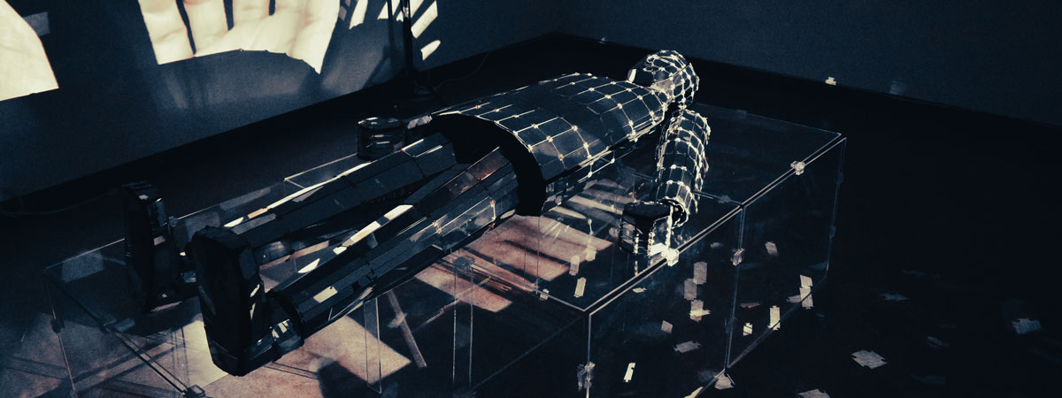 A human shape made of mirrors, emitting light, lies on a clear platform in a darkened room. Images and shapes are projected on the wall.