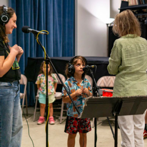 A child stands at a microphone wearing headphones, looking up at a teaching artist. An adult wearing headphones stands to the side looking on and smiling.