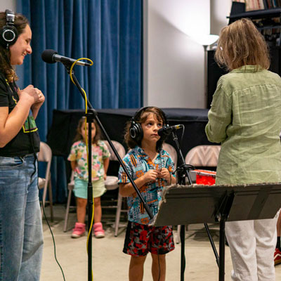 A child stands at a microphone wearing headphones, looking up at a teaching artist. An adult wearing headphones stands to the side looking on and smiling.