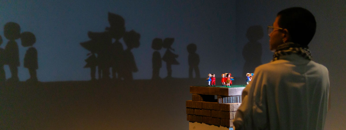 Maya Nguyen in the right foreground, with a sculptural brick platform containing several small plastic toy figures. The silhouette of those figures is shown on the wall opposite, taking up most of the image.
