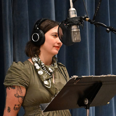 Nhatt Nichols wears headphones and stands at a microphone with a music stand in front of her.
