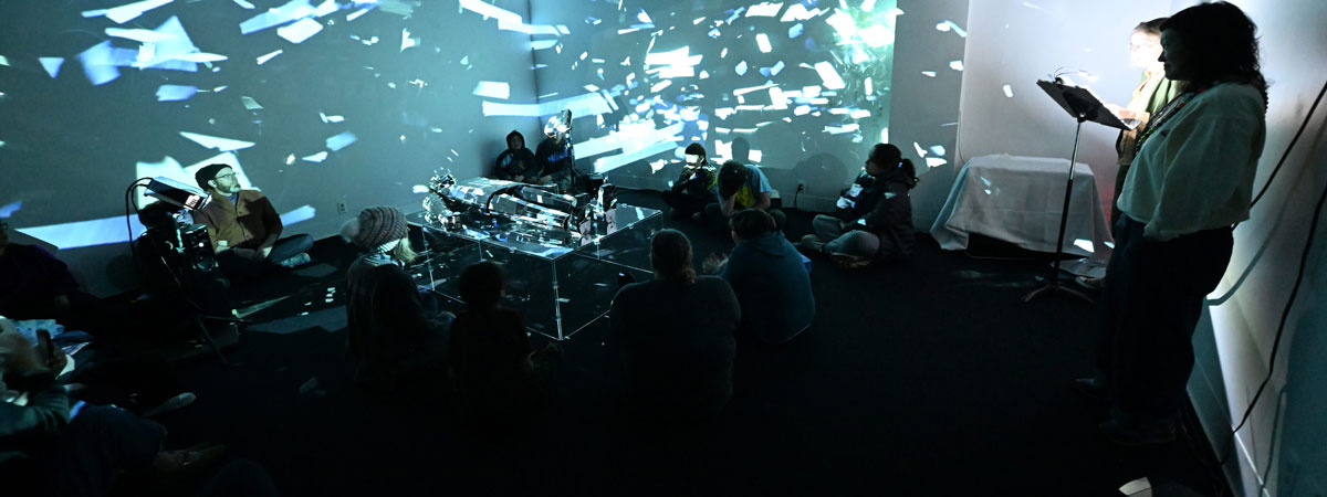 A darkened gallery space with greenish lights projected on walls. Several people sit around a sculptural object in the center. Two stand off to the right.