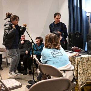 People in a recording studio. One jumps in front of a microphone, one sits at a computer, others look on.
