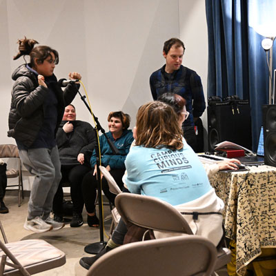 People in a recording studio. One jumps in front of a microphone, one sits at a computer, others look on.