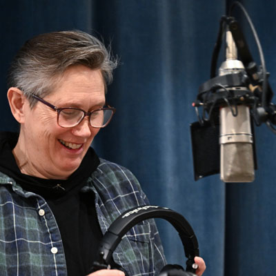 Hillary Behrman stands at a microphone, holding headphones and smiling.