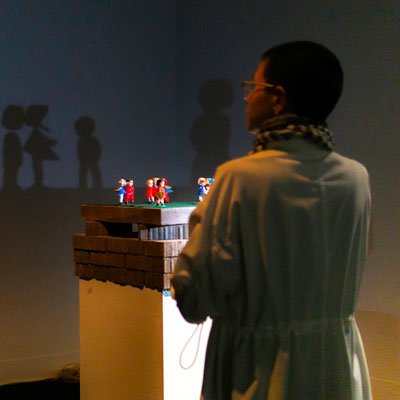 Maya Nguyen stands in the Jack Straw New Media Gallery. In front of her is a group of small figures on a pedestal. A light casts their shadows on the wall.