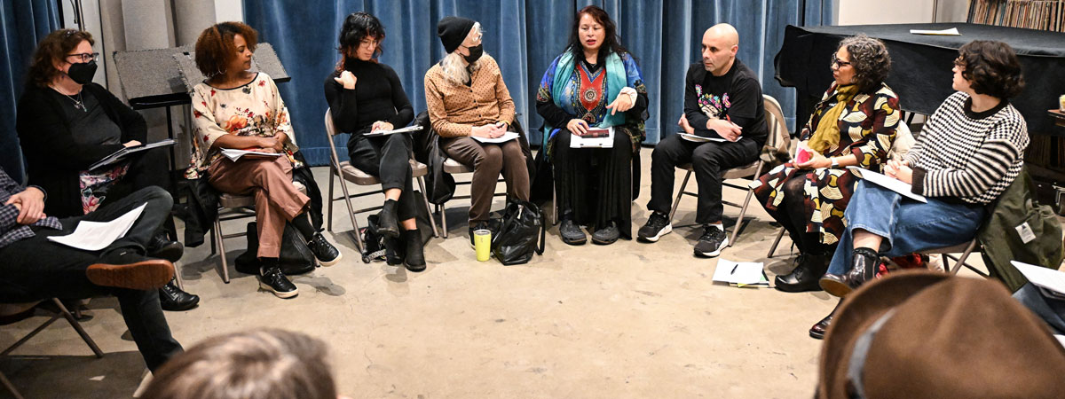 A group of people sitting in a semi-circle in a studio with concrete floors and blue curtains.