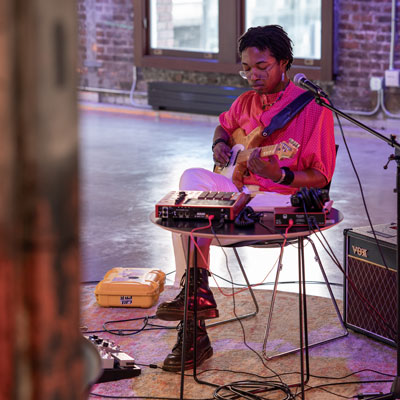 Jai sits and plays electric guitar, pedals and electronic devices on the floor and a table in front of them.