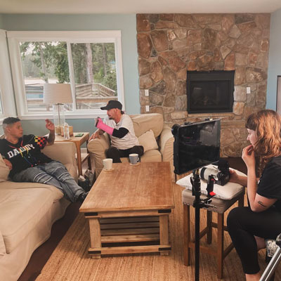 Two people sit in a living room while a third looks on, with a camera and monitor.