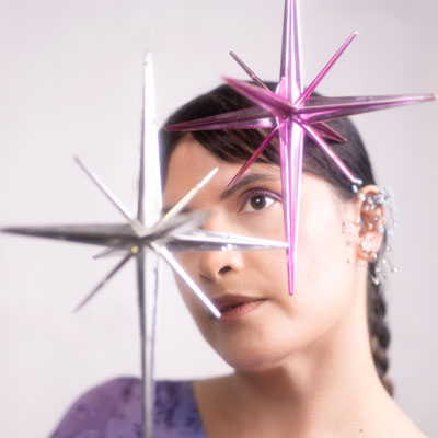 Briana Marela Lizárraga, in soft focus, with two shiny star-shaped objects floating in front of her, one silver and one purple.