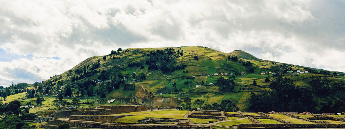 A green hill with ancient structures in the foreground. Bright, cloudy sky overhead.