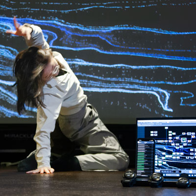 Rose Xu performs with one hand on a stage and the other in the air. A laptop computer is on the stage in front of her and an abstract image is projected behind her.