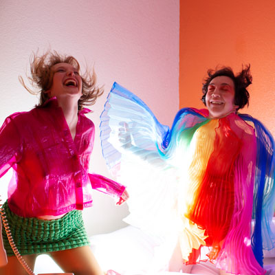 Jenny Peterson and Kaitlin McCarthy wearing brightly colored outfits, dancing in front of a pink background.