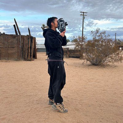 jade wong stands in a sandy landscape, facing right and looking through a 16mm film camera.