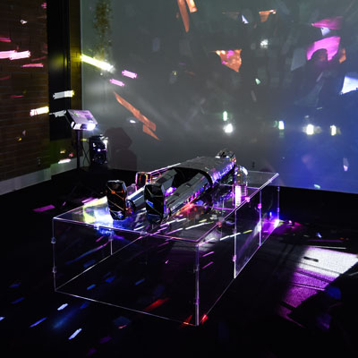 A human shape made of mirrors lies on a clear platform in a darkened room. A projector on the left points toward it. Brightly colored, fragmented shapes are projected on the wall.