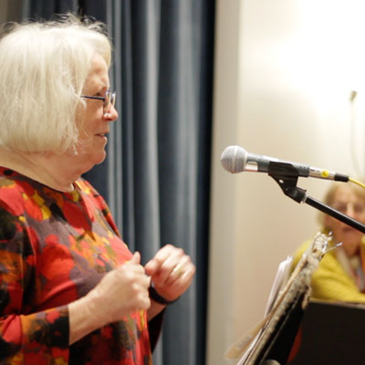 Pamela Moore Dionne stands on the left, speaking into a microphone. On the right, out of focus in the background, Lynette Westendorf sits at a piano.