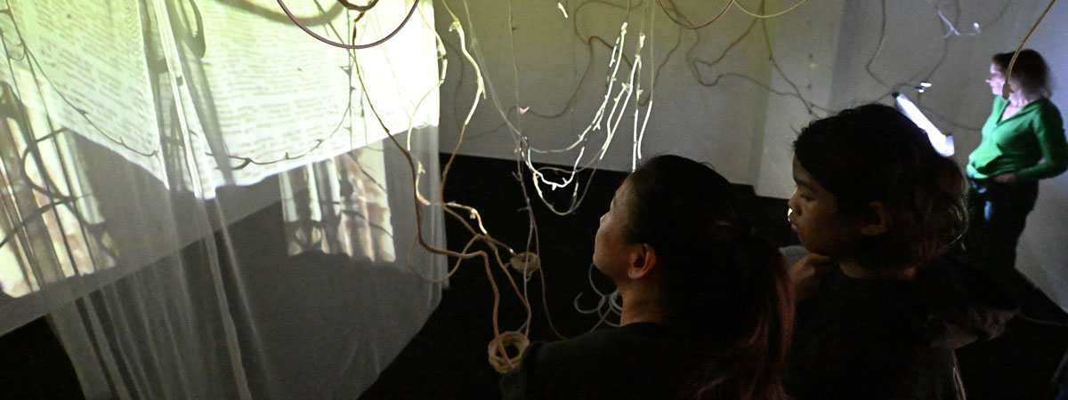 THree people in a darkened gallery space with hanging fabric, vine-like objects, and video projections.