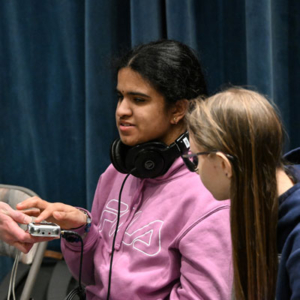 A young person wearing a pink sweatshirt and headphones has her hand on a small device the headphones are connected to. Another young person looks on.