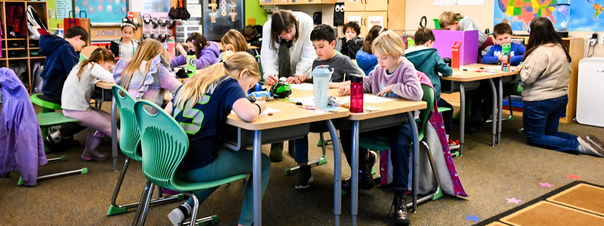 A classroom of elementary students working at their desks. In the center, Vicky stands next to one student leaning down. Another adult kneels next to a student on the right.