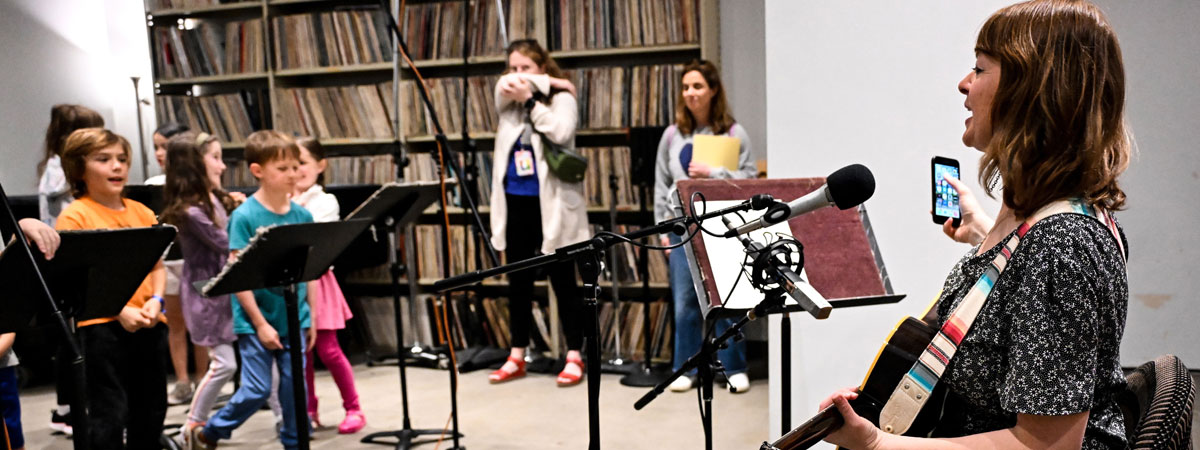 Tekla seated with a guitar in a recording studio, facing several children.