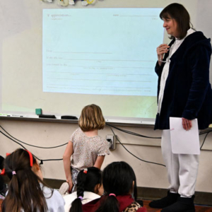 Vicky stands at a screen in a classroom showing a projection of a sheet of paper with writing on it. Students seated on the floor.