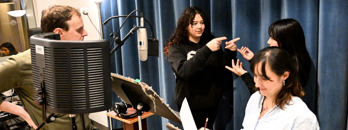 Daniel and Iveliz in a recording studio with two students near a microphone, pointing at each other.