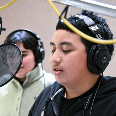 A student wearing headphones at a microphone. Stephanie, wearing headphones, in the background.