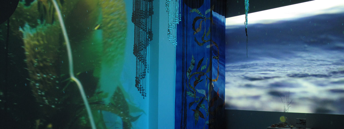 Projections on two walls show kelp in the ocean and waves. Tapestries and other objects hang from above.