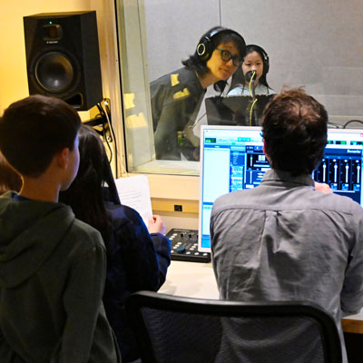 Daniel and students in a studio control room at a computer. Mimi and another student, wearing headphones, on the other side of a large window.