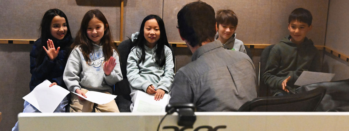 Five students holding papers, talking with Daniel in a recording studio control room.