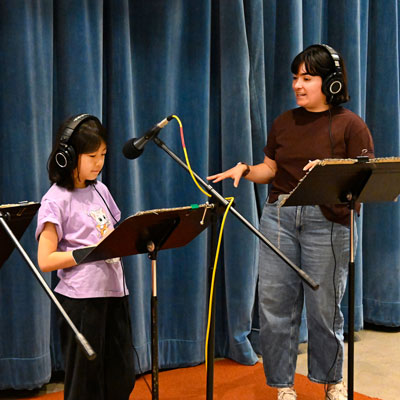 Stephanie and a student wearing headphones in a recording studio. Student stands in front of a microphone.