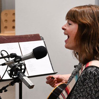 Tekla sings into a microphone, holding a guitar