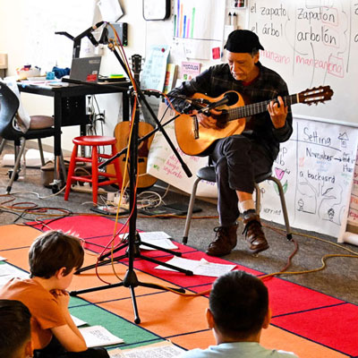 Abel sits at a microphone in a classroom playing guitar.