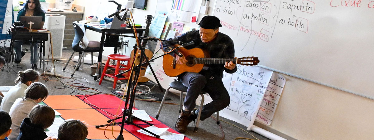 Abel, seated in a classroom, plays guitar with microphones in front of him. Ayesha sits at a computer in the background, students seated on the floor to the left.