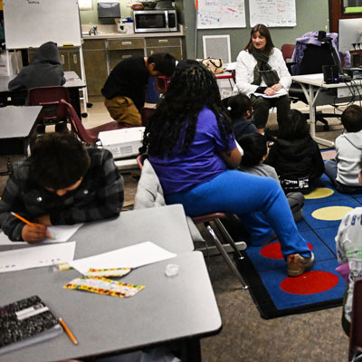 Vicky seated in a classroom with students listening to her or writing on paper with pencils.