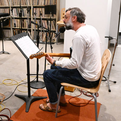 Tito sits playing electric guitar with a music stand and microphone in front of him.