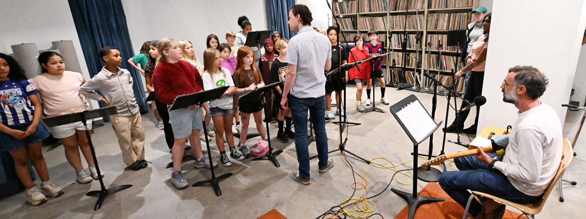 A group of students in a recording studio with music stands. Daniel stands facing them. Tito sits on the right with an electric guitar.