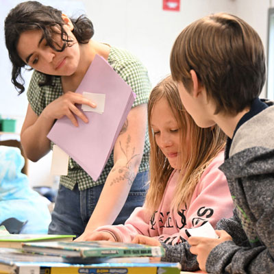 Gabriella leans over a table where two students are seated, looking at their work.